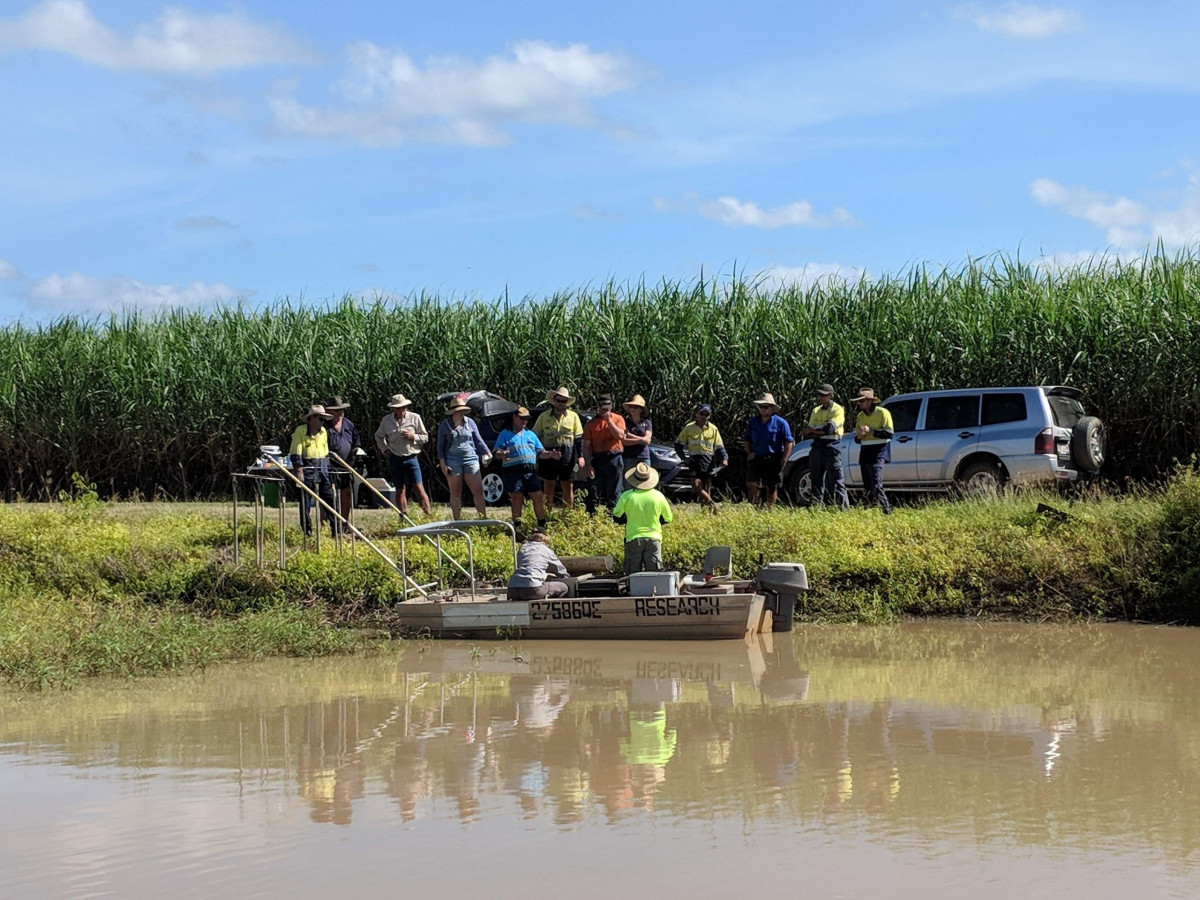 A group of people standing on the bank of a waterbody listening to researchers in a boat. Sugarcane fields in the background.