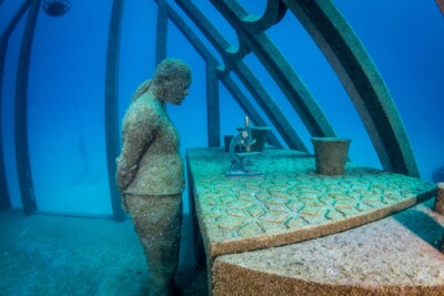 Underwater photo showing a statue of a person standing over a microscope at the Museum of Underwater Art (MOUA), John Brewer Reef.