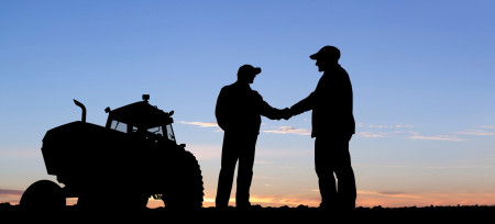 A silouette of two people shaking hands in an agricultural field with a tractor in the background.